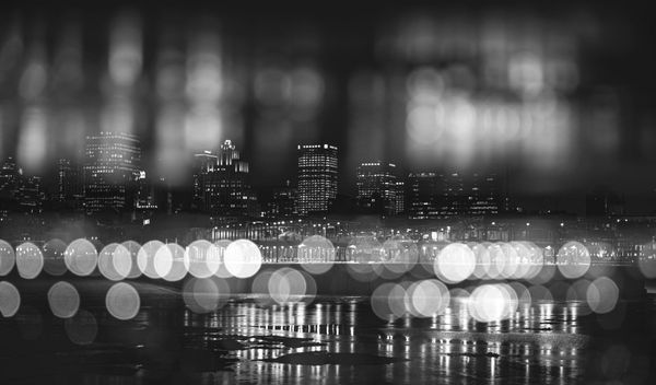 A black-and-white city skyline is blurred by large bokeh lights, creating a dreamy, abstract effect. The mood is serene and atmospheric, with reflections on water.