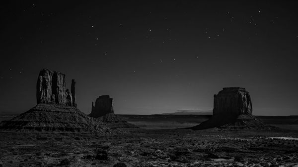 Desolate landscape of Monument Valley at night. Three silhouetted buttes under a starry sky evoke a serene and mysterious atmosphere.