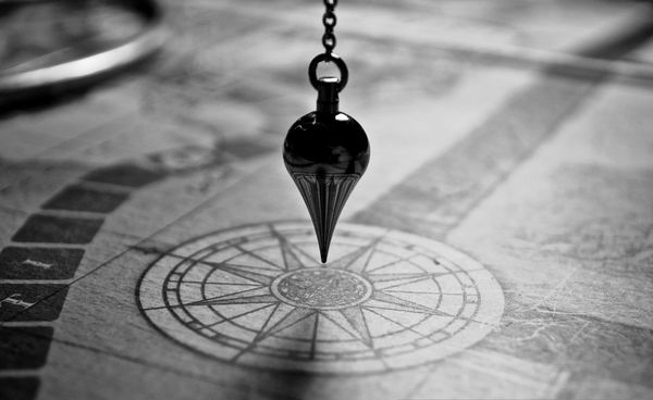 A dark pendulum hovers over a vintage map with a compass design, casting a shadow. The monochrome image evokes a sense of mystery and exploration.