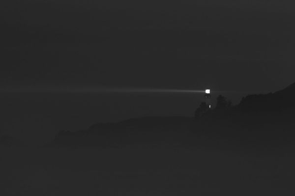 A lone lighthouse emits a bright beam across a dark, foggy landscape. The light cuts through the night, creating a sense of solitude and guidance.