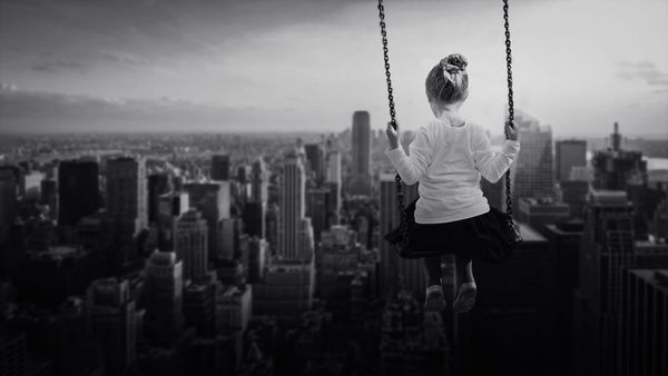 Black-and-white image of a woman on a swing, seen from behind, overlooking a dense city skyline; the scene feels quiet and contemplative.