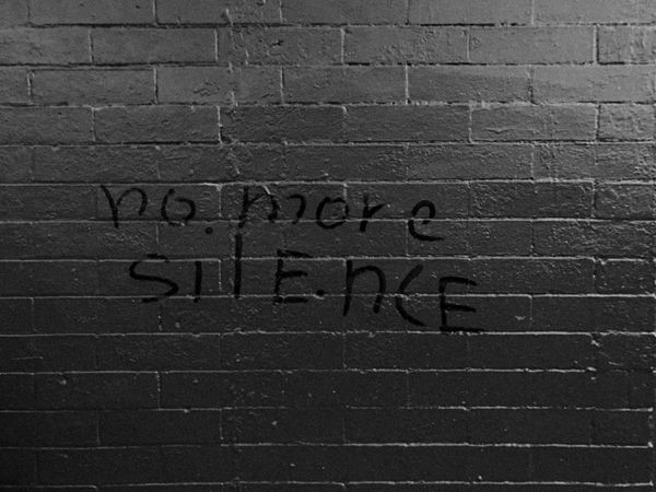 Dark brick wall with the black handwritten words “No more silence,” featuring a tall vertical stroke. The sparse, subdued scene feels somber and reflective.