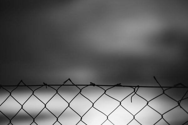 A black-and-white chain-link fence spans the foreground, its uneven top wire silhouetted against a softly blurred, hazy sky, creating a bleak mood.