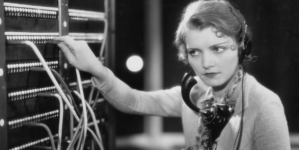 A vintage black-and-white photo of a telephone operator in a headset and mouthpiece, plugging a cord into a switchboard lined with jacks while cables hang below her hand.