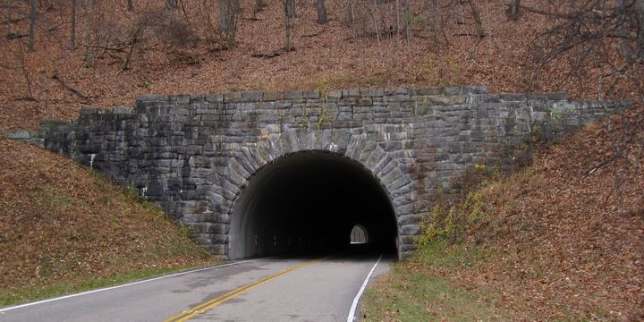 Stone tunnel entrance on the Blue Ridge Parkway, cut through a wooded hillside, with the tunnel's far end visible as a point of light.