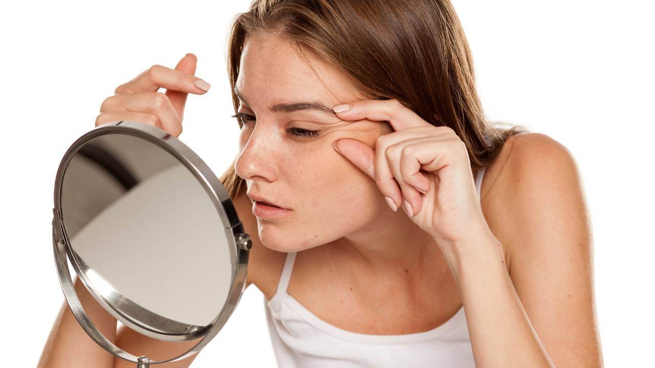 A woman pinching the delicate skin around her eye, causing dynamic wrinkles and fine lines while looking into a mirror.