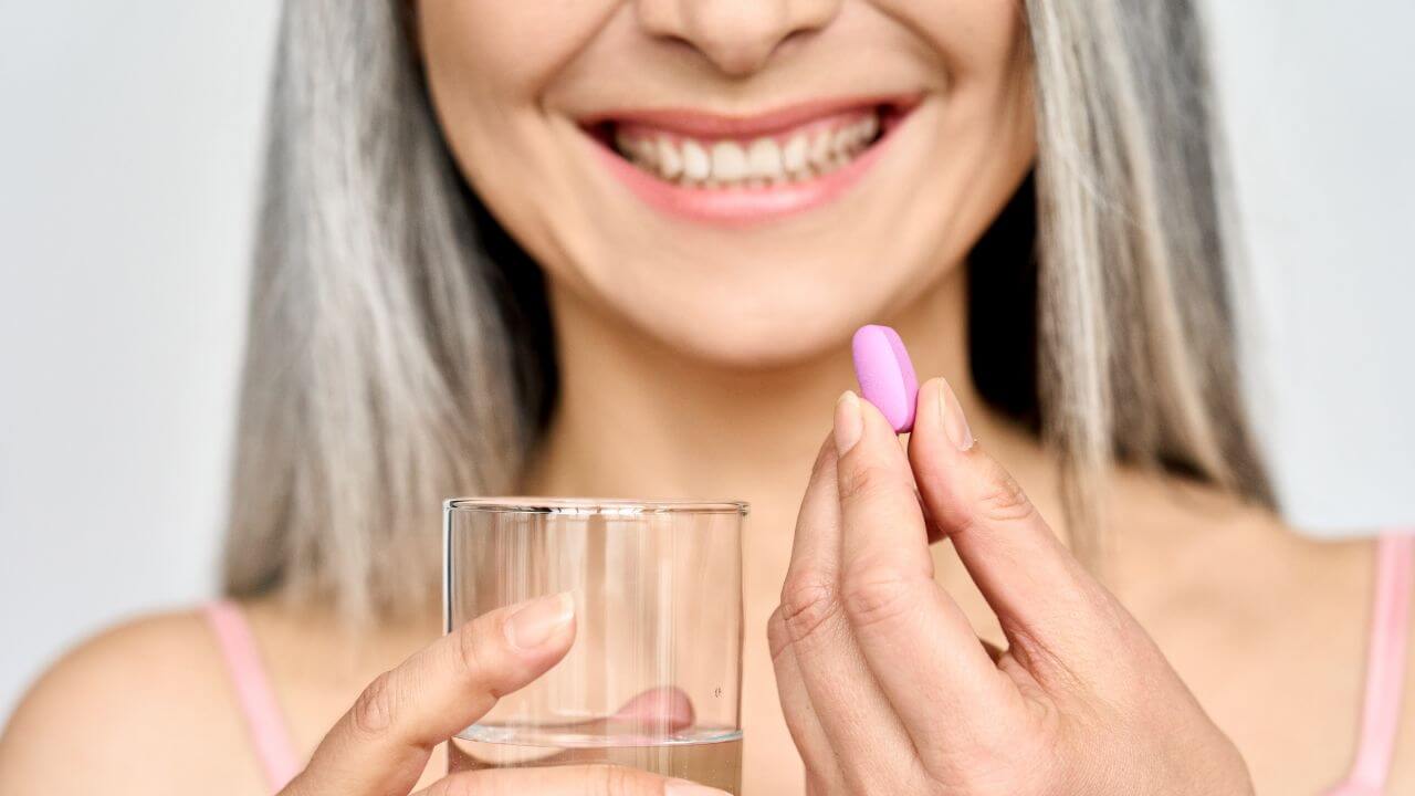 Smiling woman with gray hair holding a glass of water while taking a collagen supplement in a convenient capsule form.