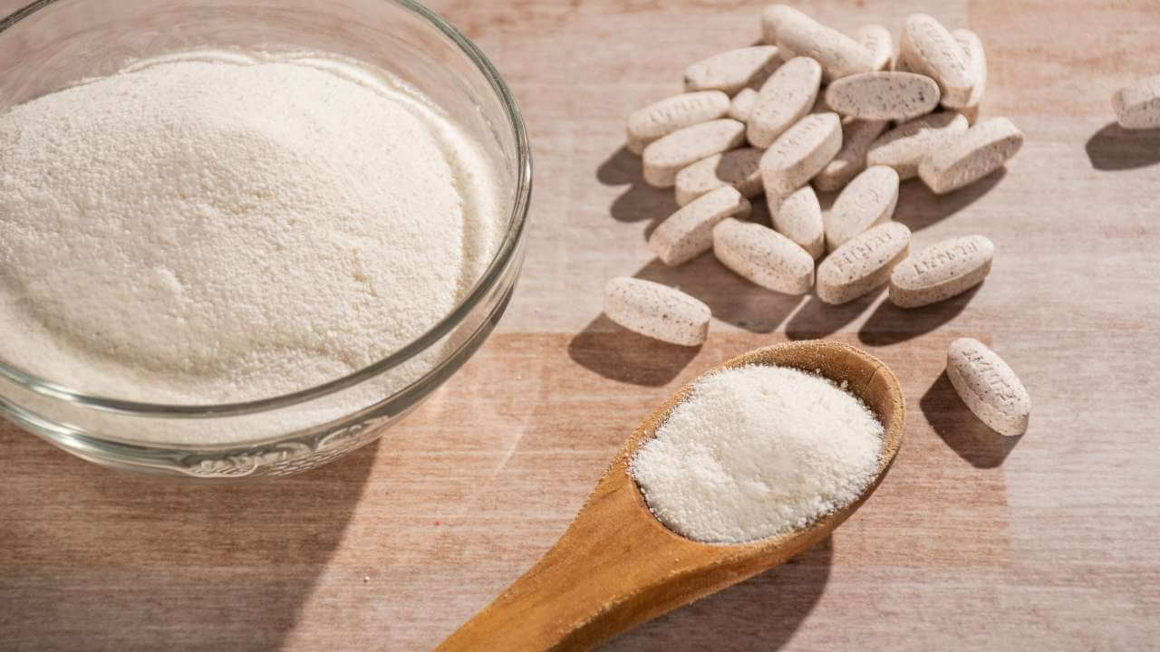 Glass bowl and wooden spoon full of collagen powder on a table next to tablets of amino acids.