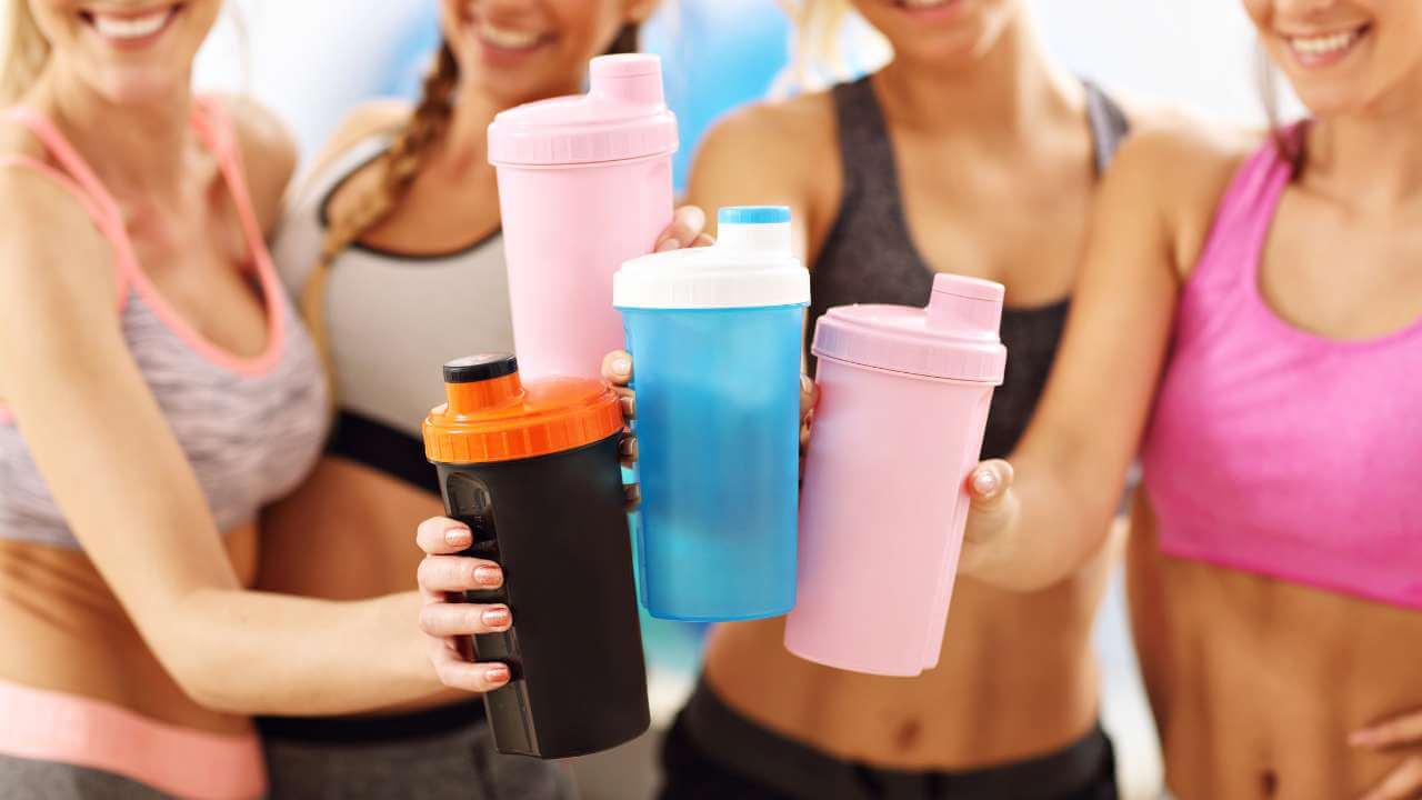 4 smiling women in workout gear toasting each other with shaker bottles of collagen powder after resistance training.