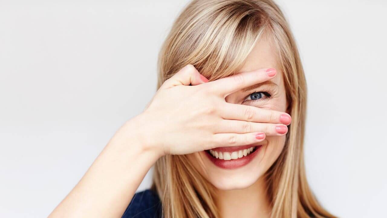 Woman’s hand covering one eye while the other peeks through her finers while making a facial expression causing crow’s feet. 