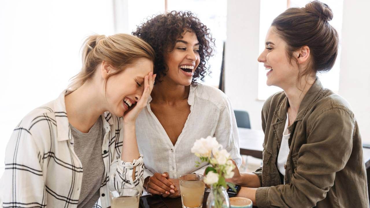 3 women laughing while having drinks at a small table.