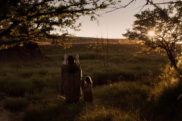 Image from Killers of the Flower Moon. An Osage woman stands with her hands up, palms facing the rays of the setting sun.