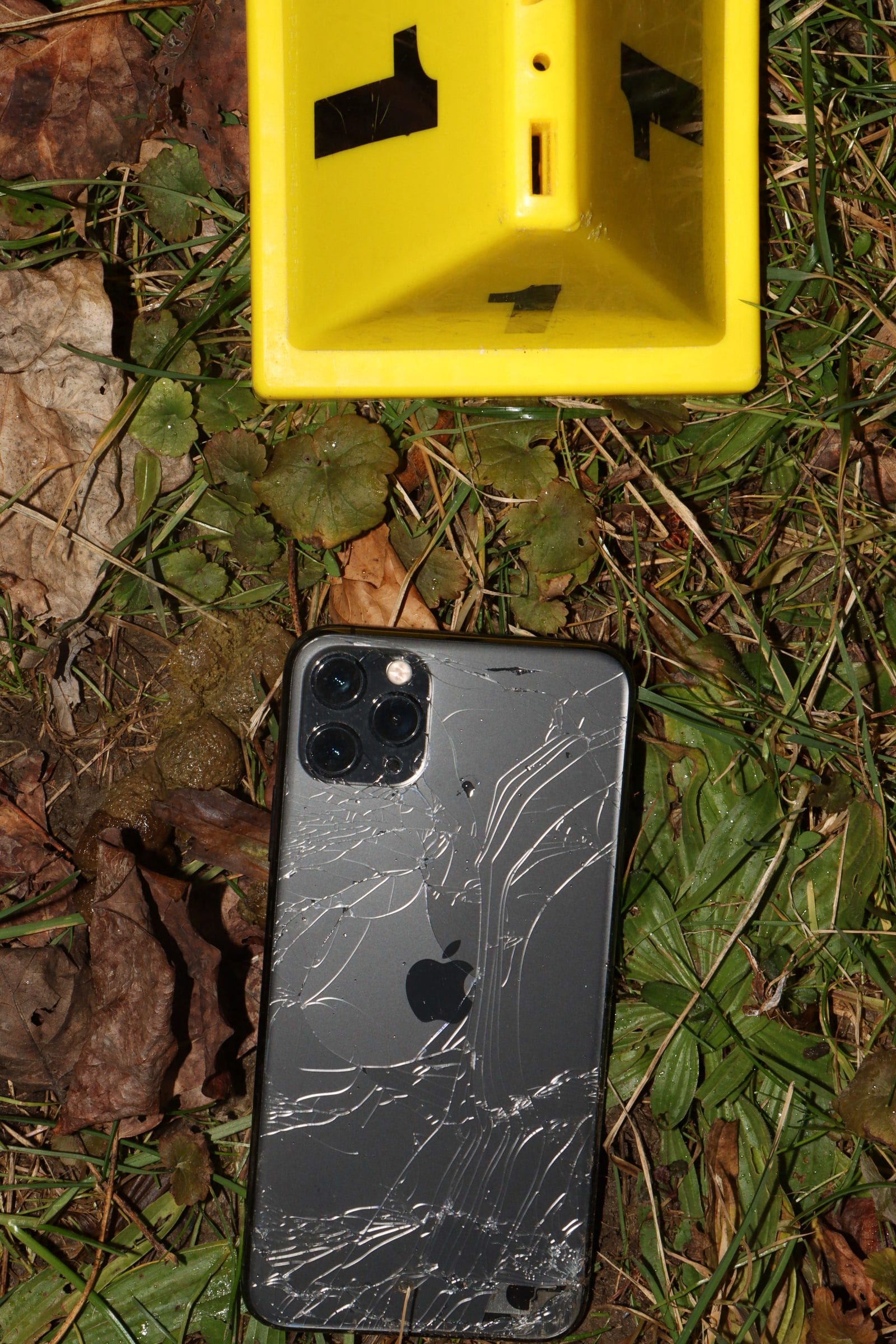 A shattered iPhone lying face-down in wet leaves and grass beside a yellow evidence marker.