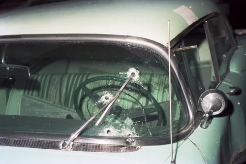 Four bullet holes through the driver's side of the windshield of a light green 1956 Buick, cracks radiating from each, the steering wheel visible behind the glass.