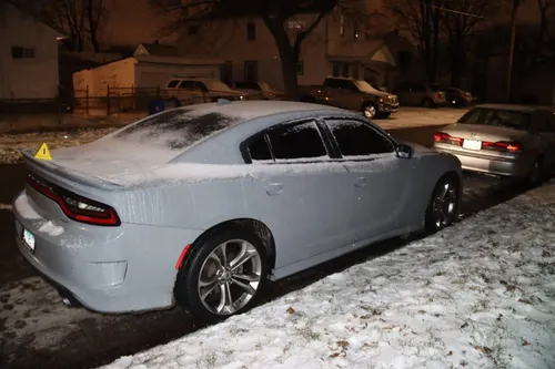 A grey Dodge Charger under fresh snow on a residential street at night, a numbered evidence marker on the boot lid.