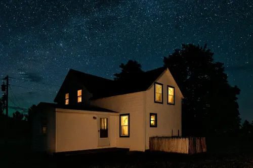 A farmhouse at night, windows lit, under a starry sky.