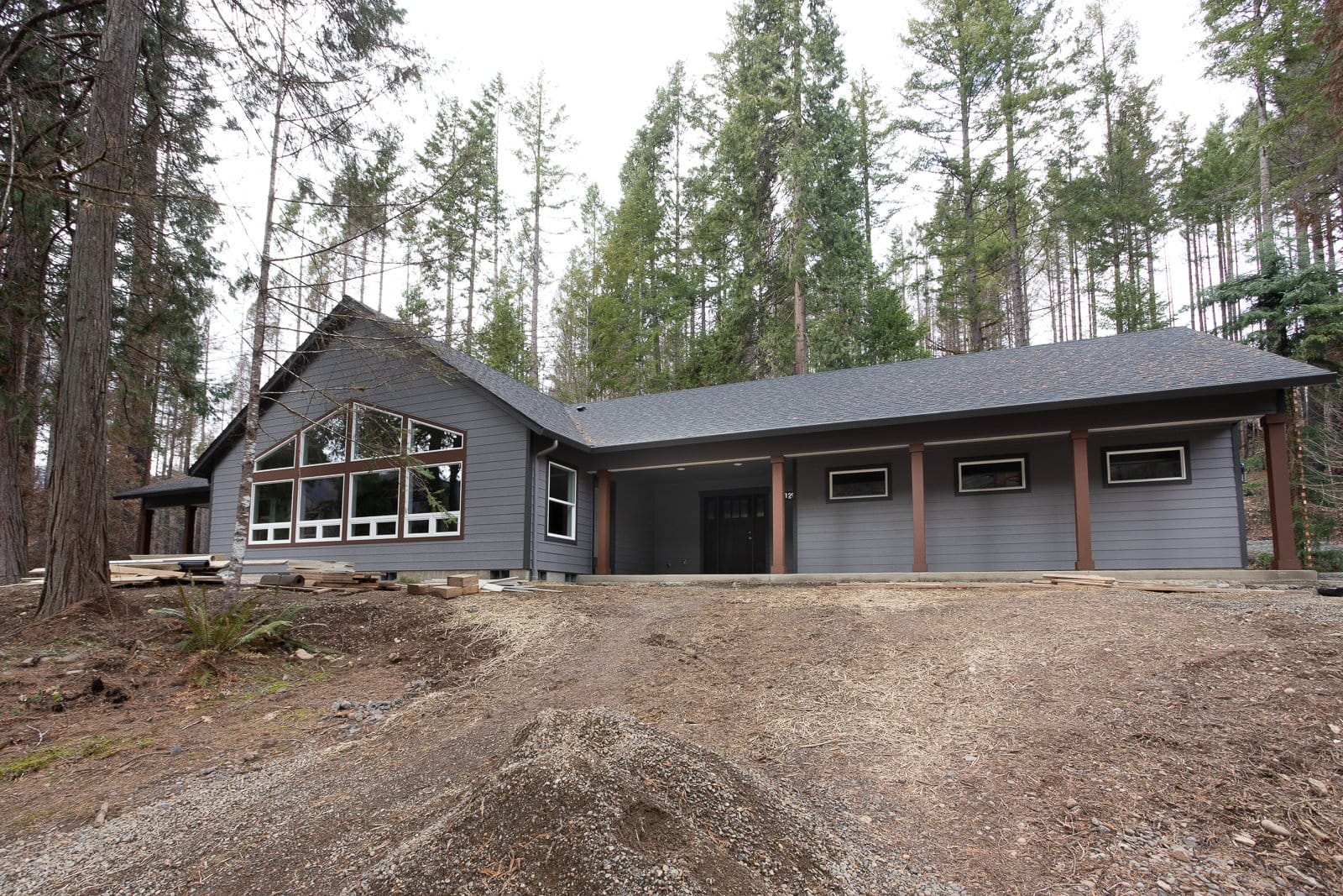 A dark gray custom home set among tall evergreen trees, featuring large front windows, a long covered porch, and a sloped lot leading to the house.