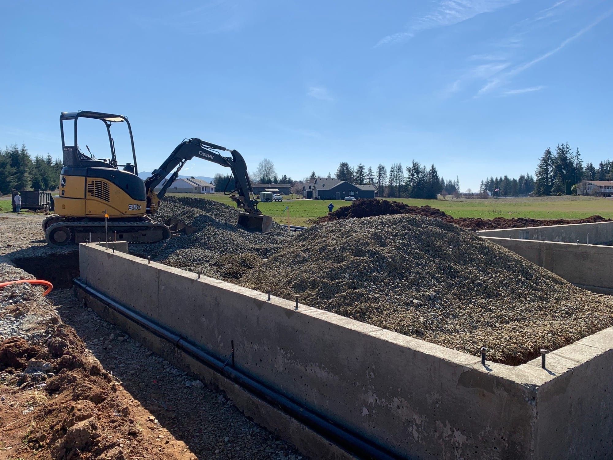 A construction site with an excavator moving gravel near a newly poured foundation, surrounded by open grassy land and nearby homes.