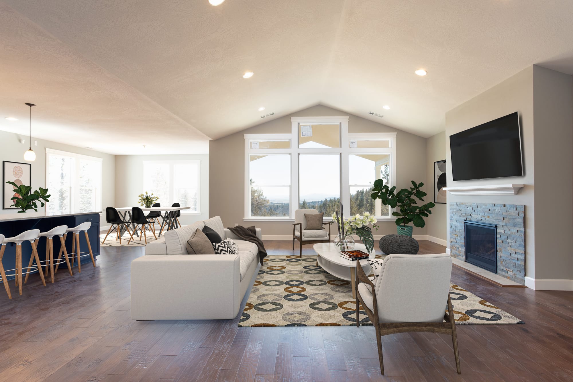 A spacious open concept living area with a cozy seating arrangement, tall windows overlooking a scenic landscape, hardwood floors, and a stone fireplace beneath a mounted TV.