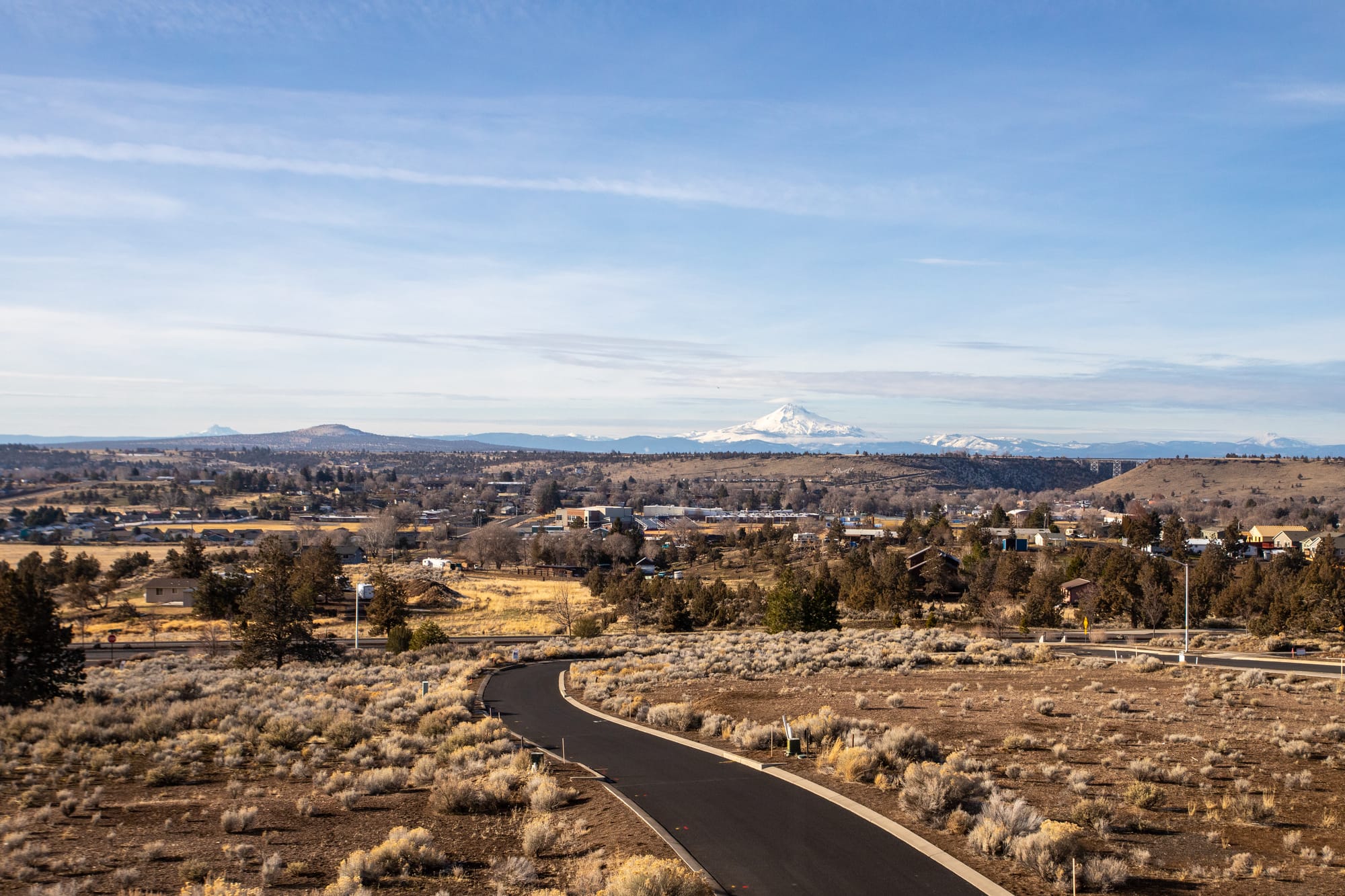 A wide landscape view of Central Oregon with open fields, scattered homes, winding roads, and snow capped mountains in the distance under a clear sky.