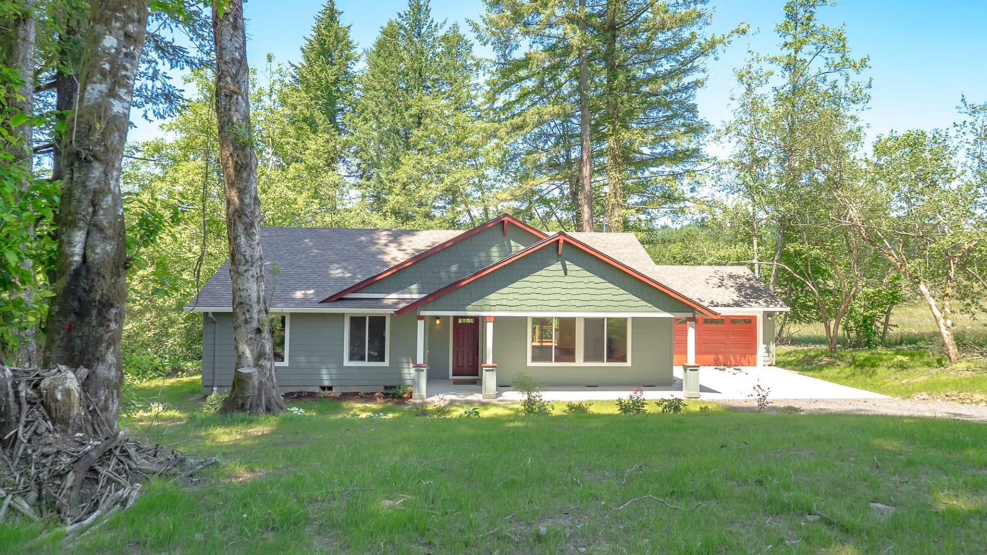 A green and red trimmed ranch style home nestled among tall trees with a grassy front yard and natural forest surroundings.