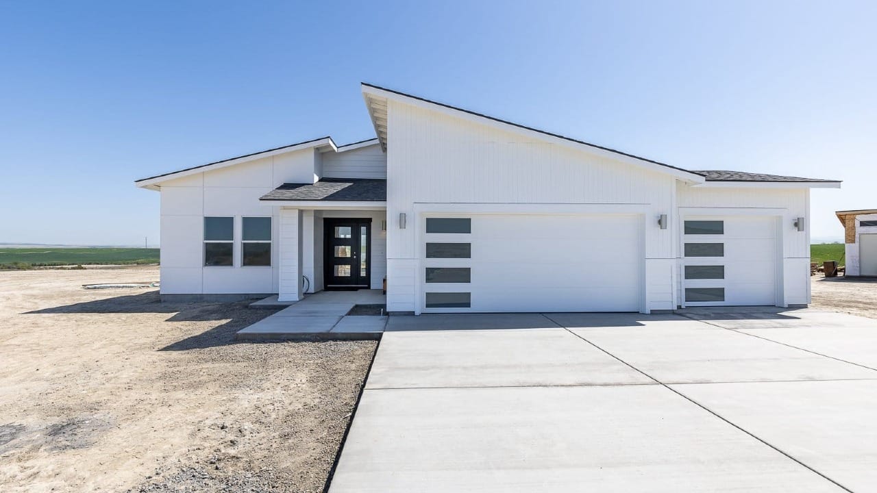A modern single-story white custom home with a large three-car garage, clean horizontal rooflines, and contemporary glass-panel garage doors, set on a wide lot with open landscape in the background.