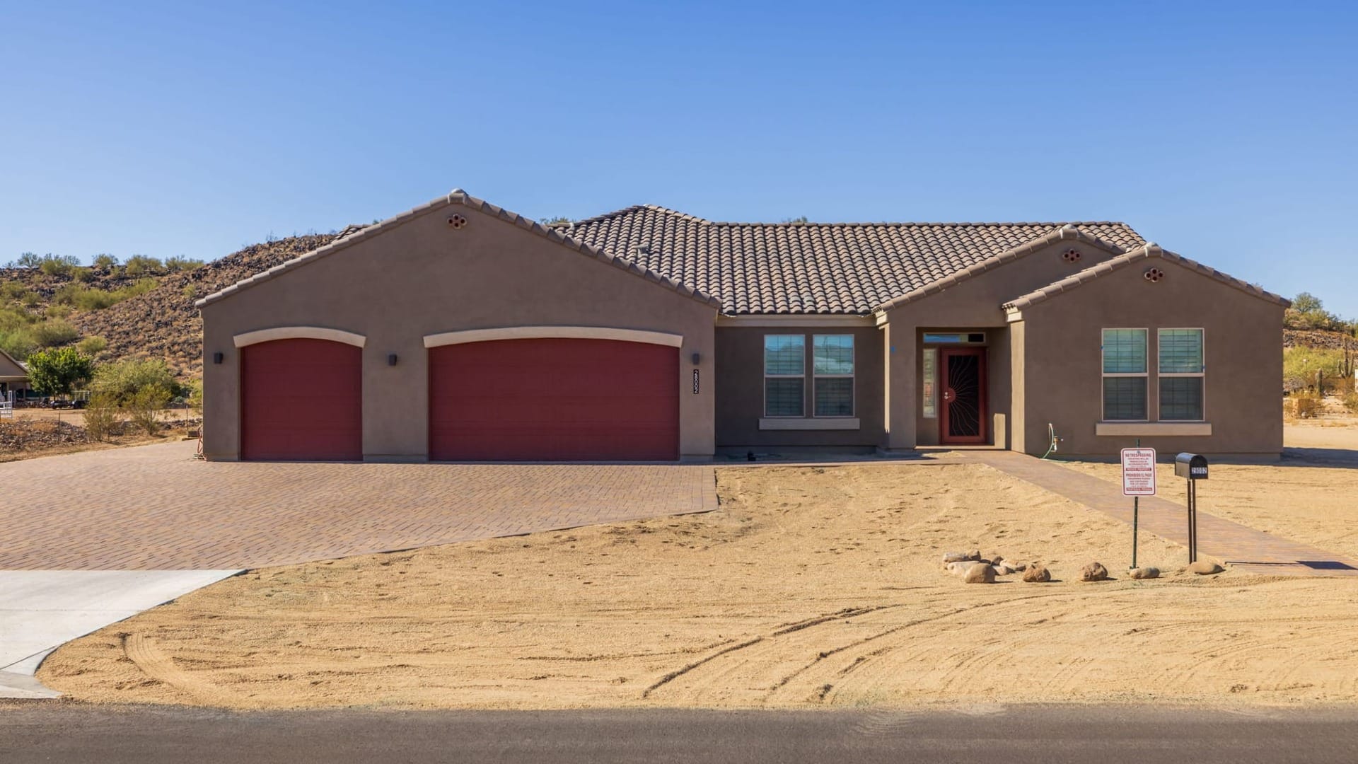 Custom built stucco home with tile roof, red garage doors, covered entry, and desert lot landscaping under a clear sky.