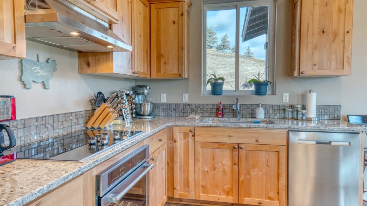 A cozy kitchen with natural wood cabinets, stainless steel appliances, granite countertops, and a window overlooking a hillside landscape.