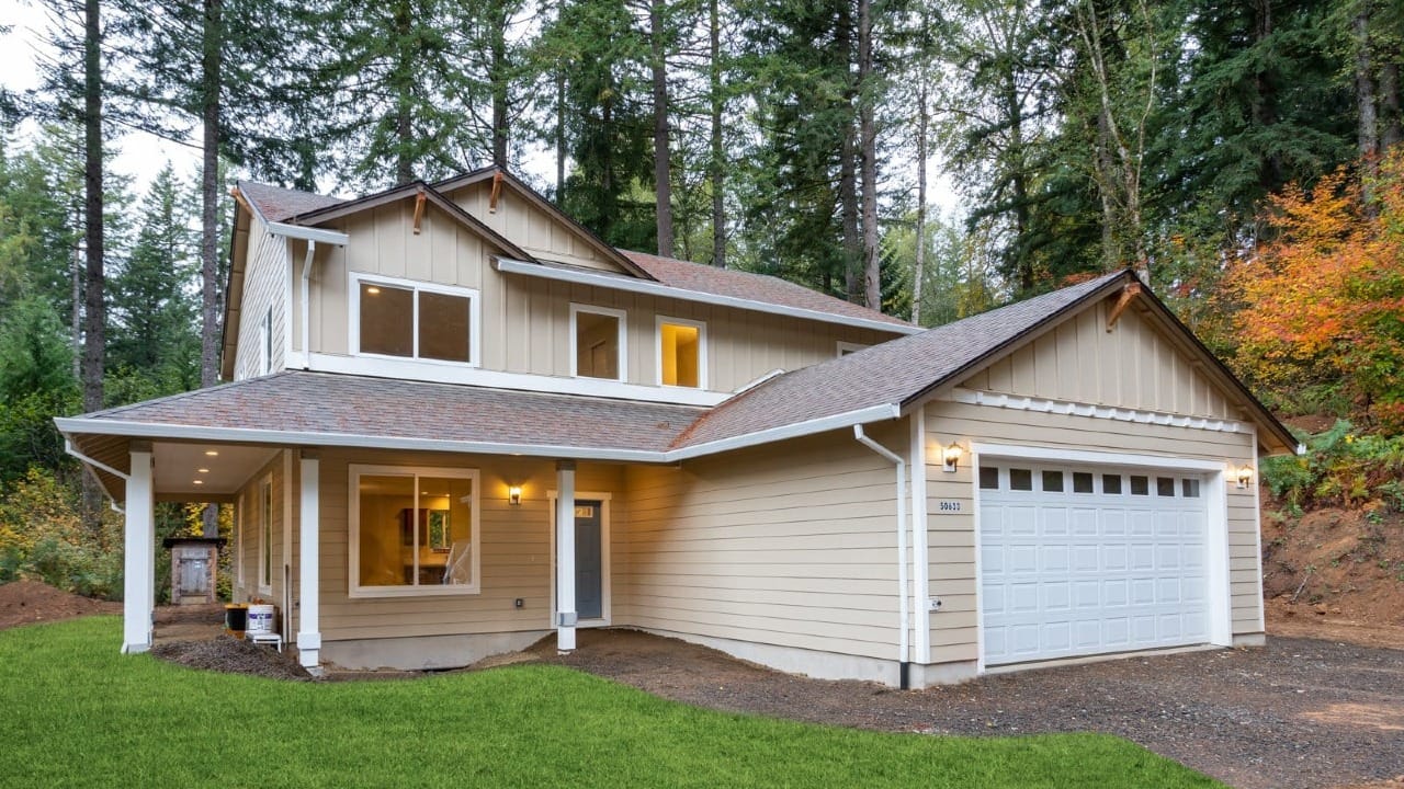 A two story beige custom home with a covered front porch and attached garage, surrounded by tall evergreen trees and a natural forest landscape.