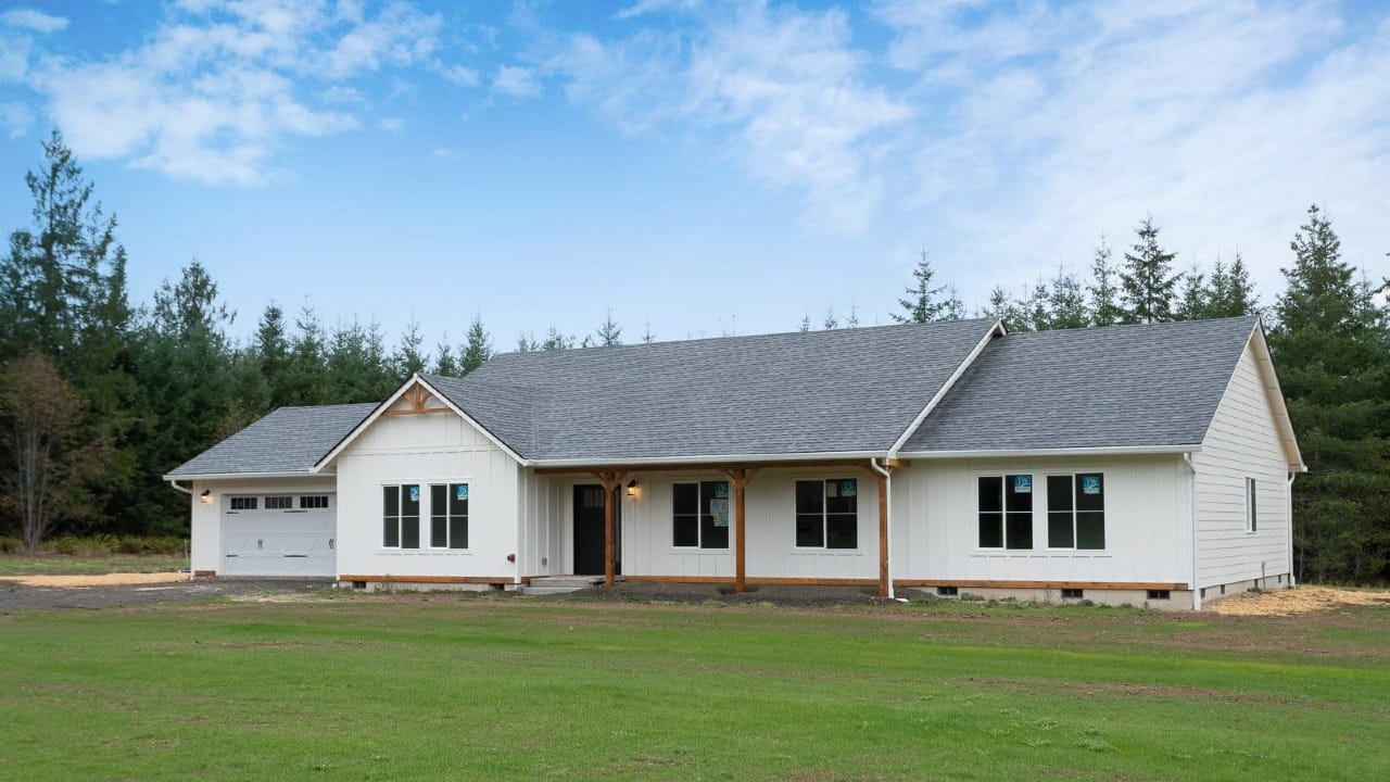 A white farmhouse style ranch home with a covered porch, two car garage, and a green lawn backed by dense evergreen trees.
