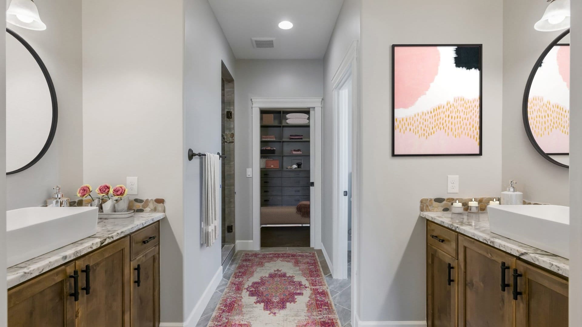 A modern bathroom with dual vanities, vessel sinks, stone countertops, and a hallway leading to a walk in closet.