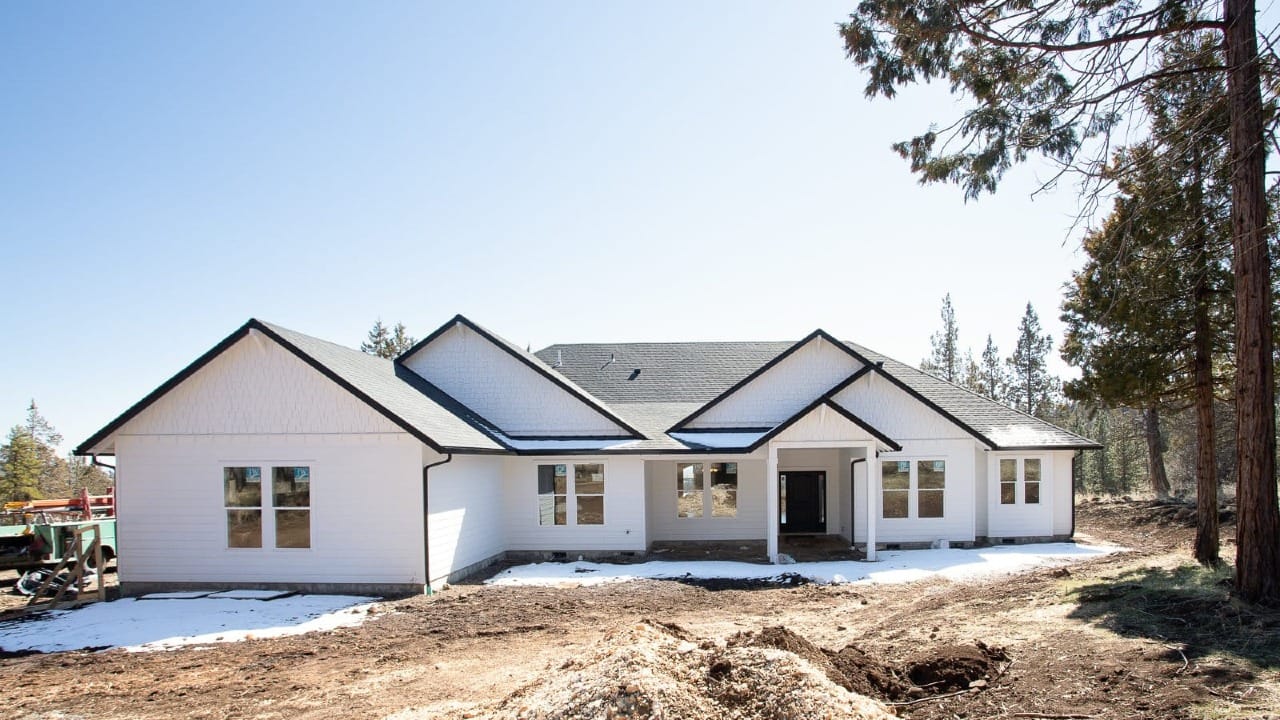 A large white ranch style custom home under construction, surrounded by dirt and tall trees in the background.