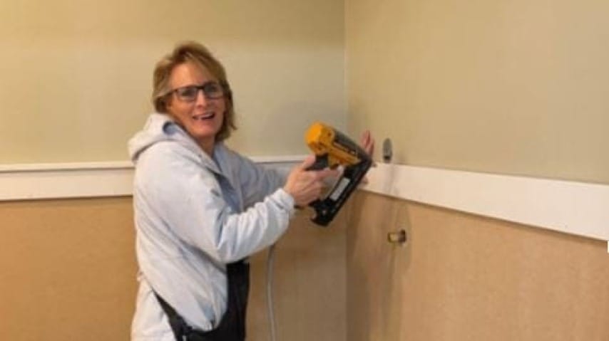 Lady using a nail gun on a wall and looking at the camera