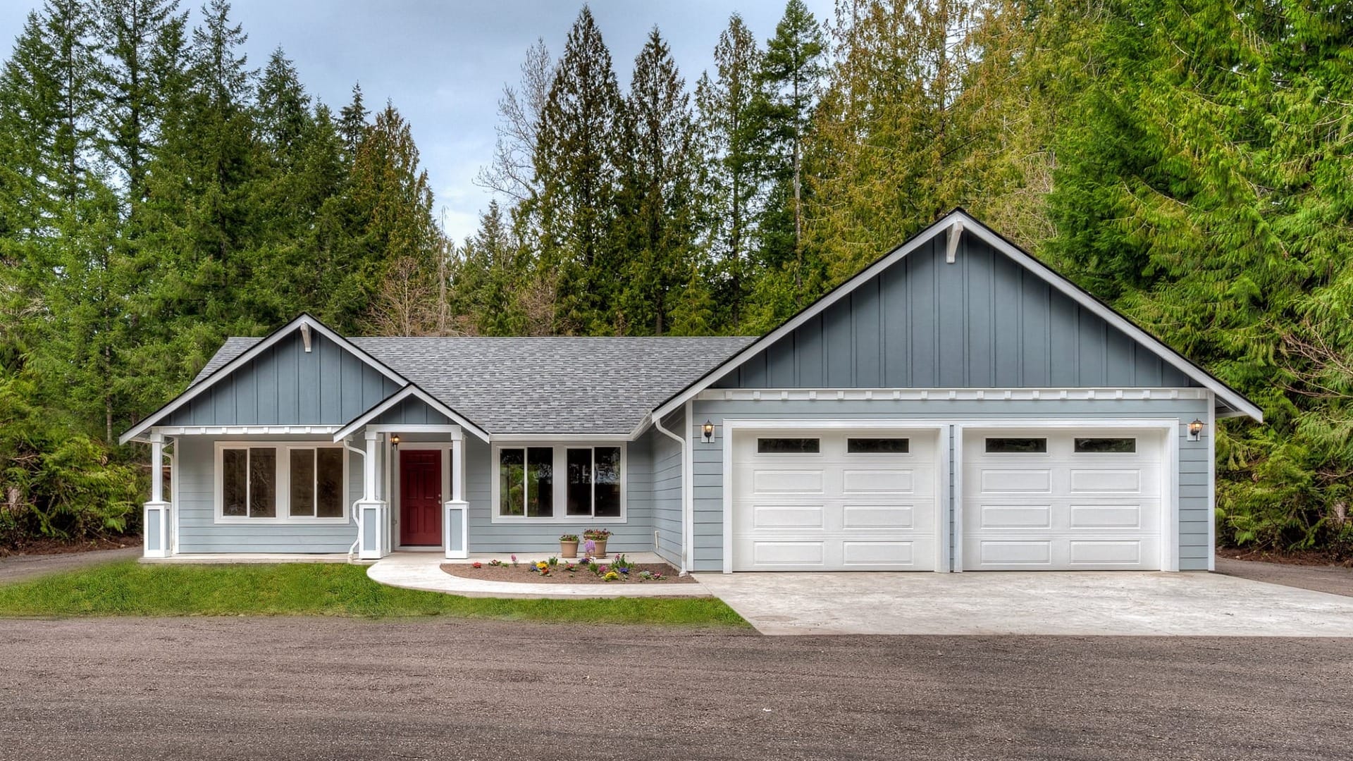 A blue ranch style home with white trim, two car garage, and a landscaped front yard set against a backdrop of tall evergreen trees.