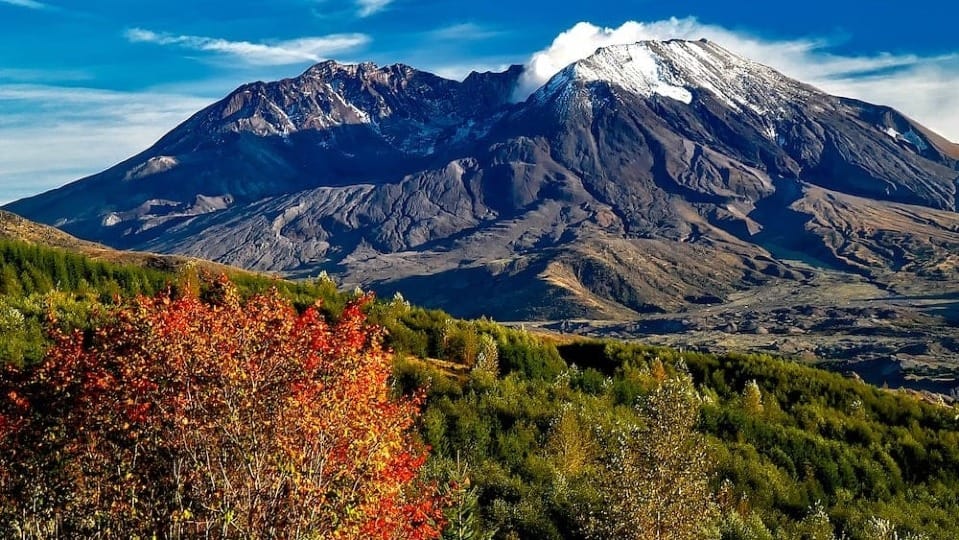 Snow capped mountain rising above forested hills with autumn colors in the foreground and clear blue sky.