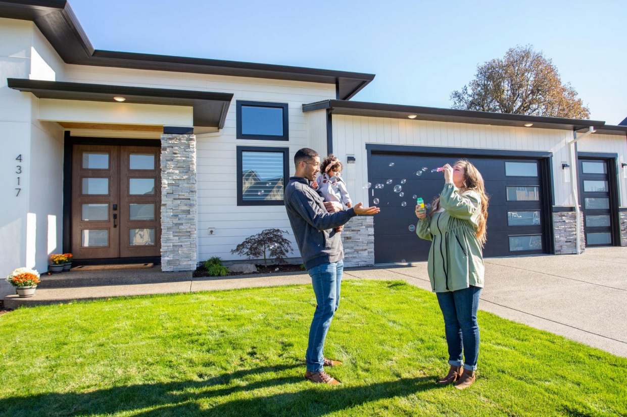 A family playing with bubbles on the front lawn of a modern white home with stone accents and dark garage doors.