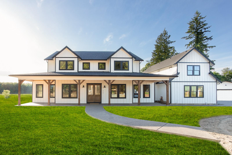 A two story modern farmhouse with white siding, black trim, a large covered porch, and landscaped green lawn under a clear sky.