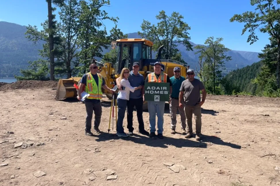 Adair Homes crew standing proudly on a construction site with heavy equipment and scenic forested mountains behind them.