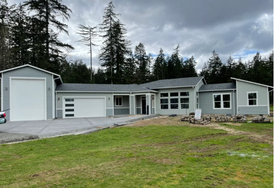 Custom single-level home with attached RV garage and traditional two-car garage, featuring light gray siding and a spacious driveway surrounded by forest.