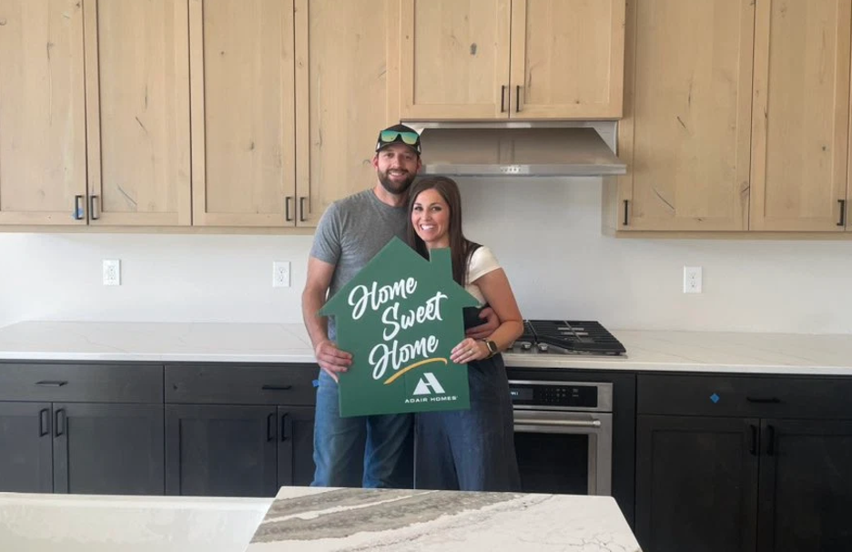 Smiling couple standing inside their new Adair Homes kitchen holding a green ‘Home Sweet Home’ sign in front of modern cabinetry and stainless appliances.