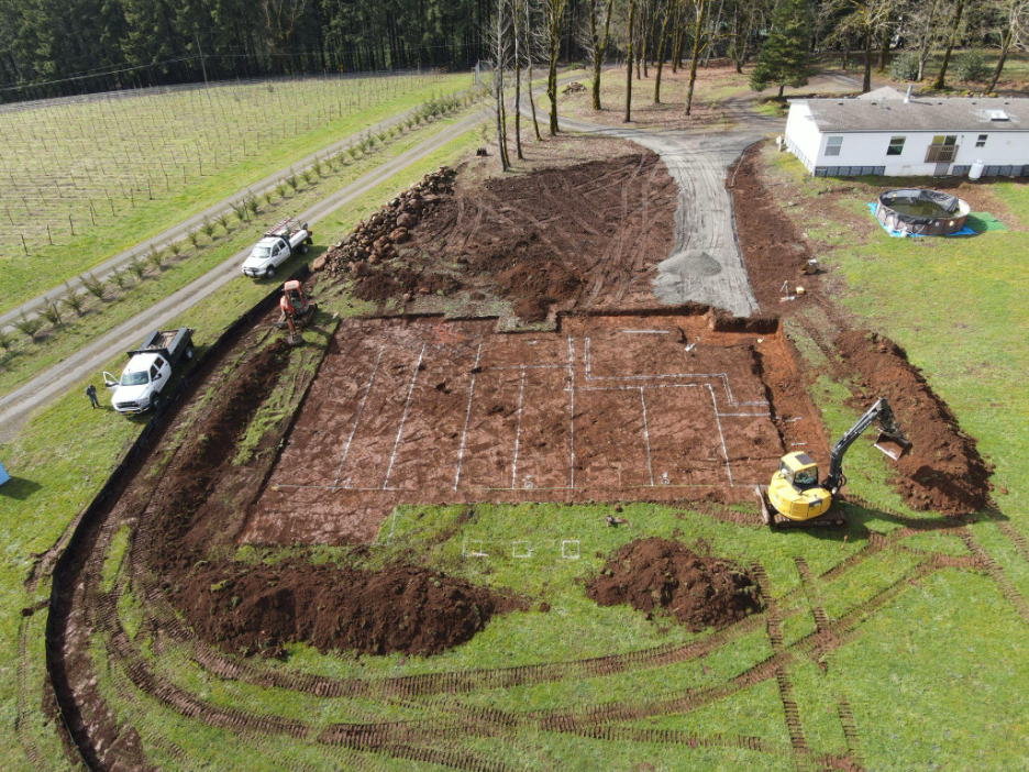 Aerial view of a residential construction site with excavation equipment and foundation layout marked in white on red soil, surrounded by farmland and trees.