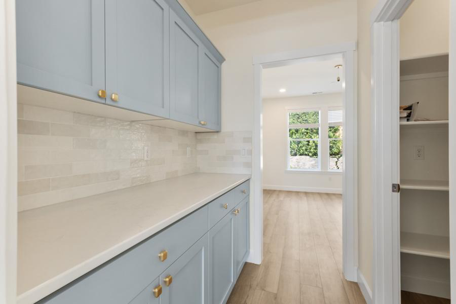 Bright butler’s pantry with soft blue cabinets, gold hardware, light subway tile backsplash, and wood flooring leading into a naturally lit dining area.