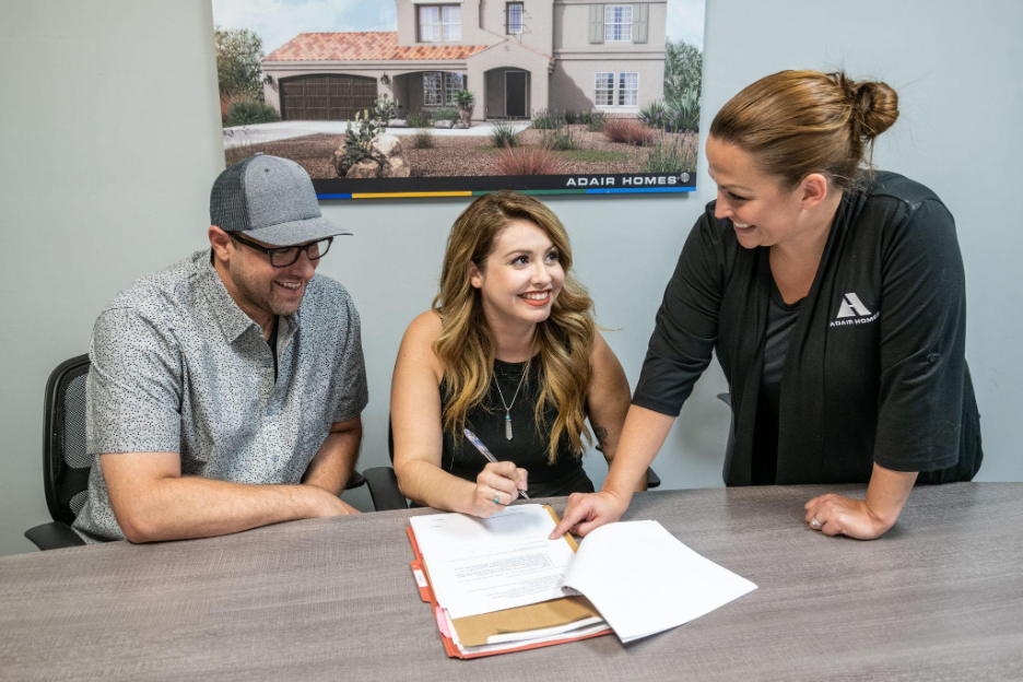 Happy couple signing their custom home agreement with an Adair Homes representative, inside a design center with floor plans and renderings displayed.