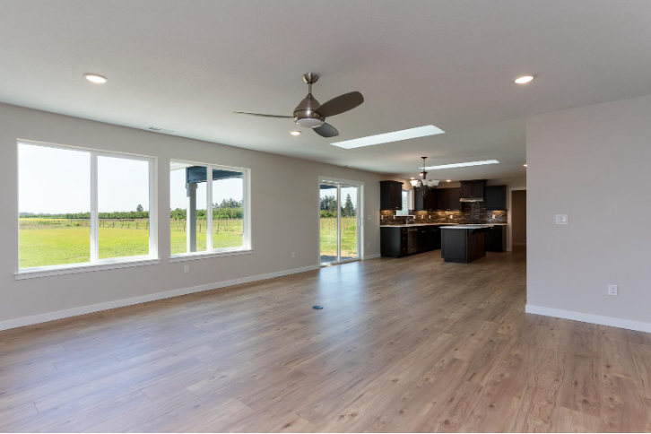 Large open great room with wood flooring, ceiling fan, skylights, and kitchen visible in the background.