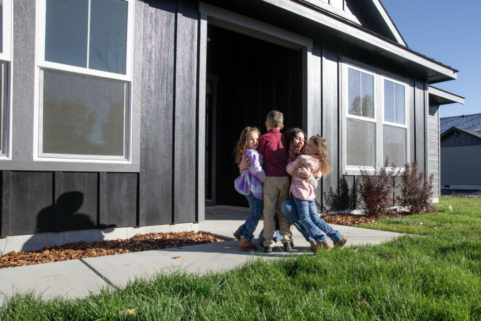 Family with young children joyfully embracing outside the front entrance of their newly built modern home.