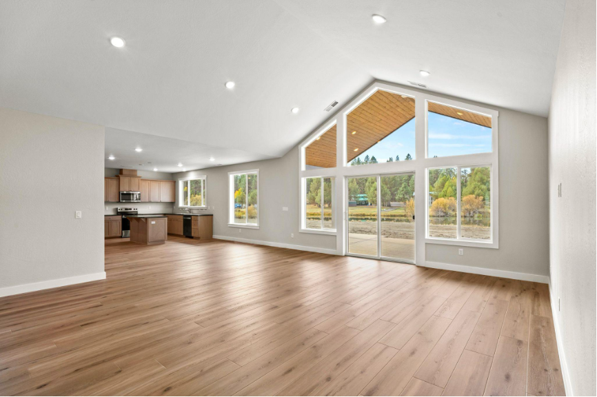 Bright open concept living area with large windows, vaulted ceiling, wood flooring, and modern kitchen in the background.