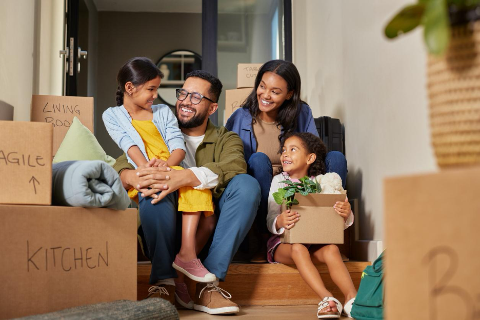 Smiling family sitting on the front steps of their new home surrounded by moving boxes on move in day.