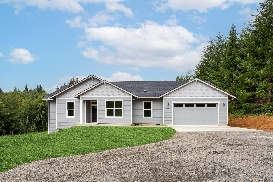 Newly built gray single story home with attached garage and forested backdrop under a blue sky.