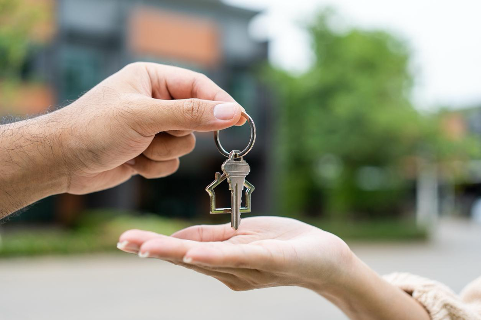 Close up of a hand placing house keys into another person’s palm in front of a blurred home exterior.