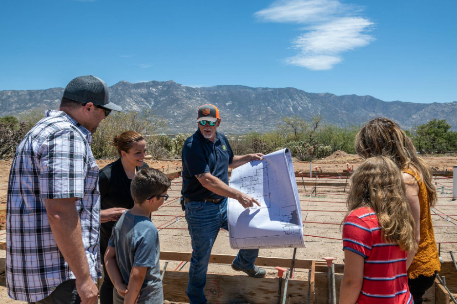 Construction superintendent reviewing blueprints with a family at a residential build site with mountains in the background.